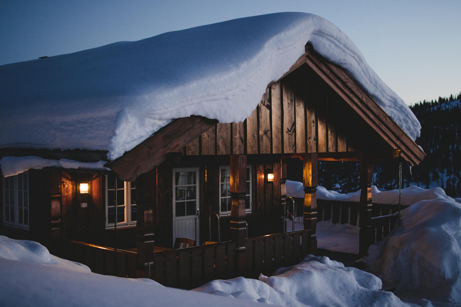 A snow-covered wood cabin illuminated warmly against a winter dusk sky, surrounded by snowy landscape.